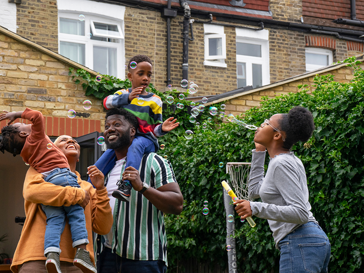 A family of standing in the garden. One girl is blowing bubbles and the dad is holding a young child on his shoulders whilst the mum is carrying another child. Both children are trying to grab the bubbles and everyone is laughing and smiling.