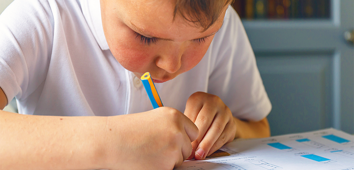 Young boy dressed in school uniform sitting in a classroom at a desk writing on a piece of paper