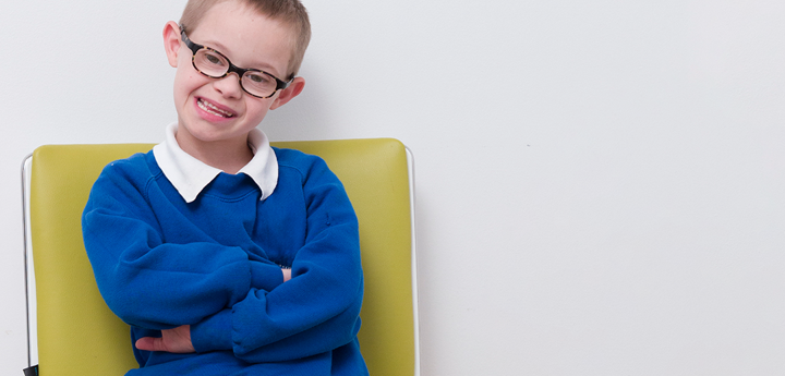 Child with Downs syndrome wearing glasses smiling. He is sitting on green chair with arms crossed.