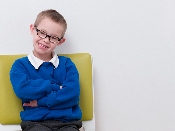 Child with Downs syndrome wearing glasses smiling. He is sitting on green chair with arms crossed.