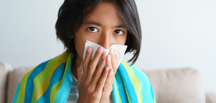 Girl sitting on sofa wrapped in a blanket, blowing nose into a tissue.