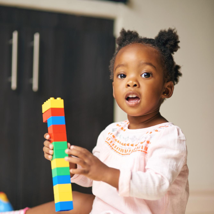 Toddler girl in pink dress sitting holding a tower of building blocks in her hand