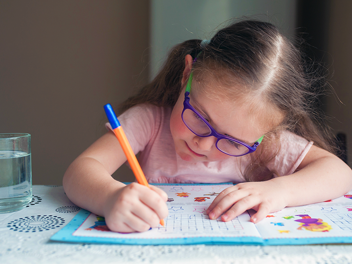 Girl Writing In Activity Book Wearing Glasses