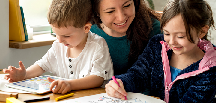 Adult with two children, one boy and one girl. The boy is sat on the adult's lap playing on a tablet. The girl is drawing a picture.