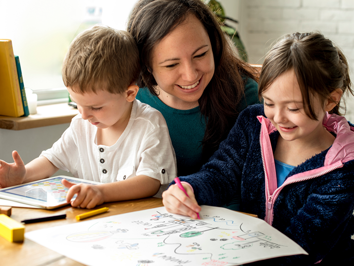Adult with two children, one boy and one girl. The boy is sat on the adult's lap playing on a tablet. The girl is drawing a picture.
