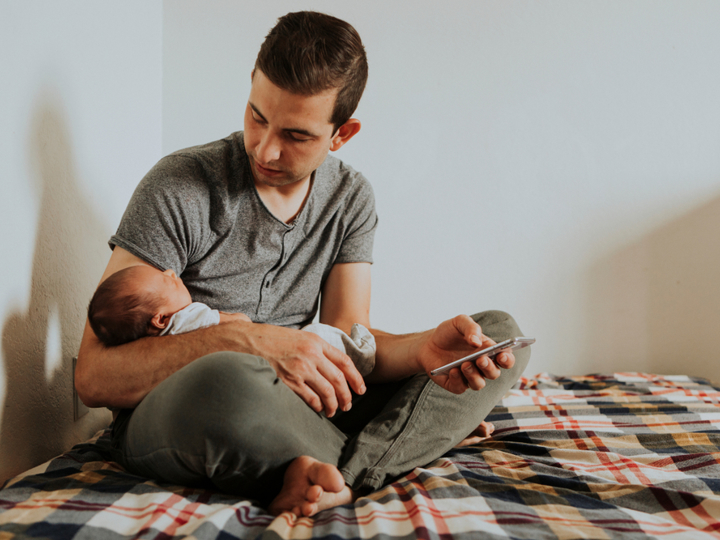 Dad sitting on bed looking down at his baby which he is holding in his arm. His other hand is holding his phone.