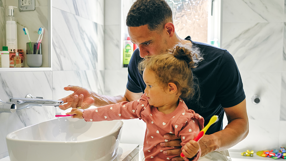 Child holding a toothbrush under a tap while sitting on adult's lap.