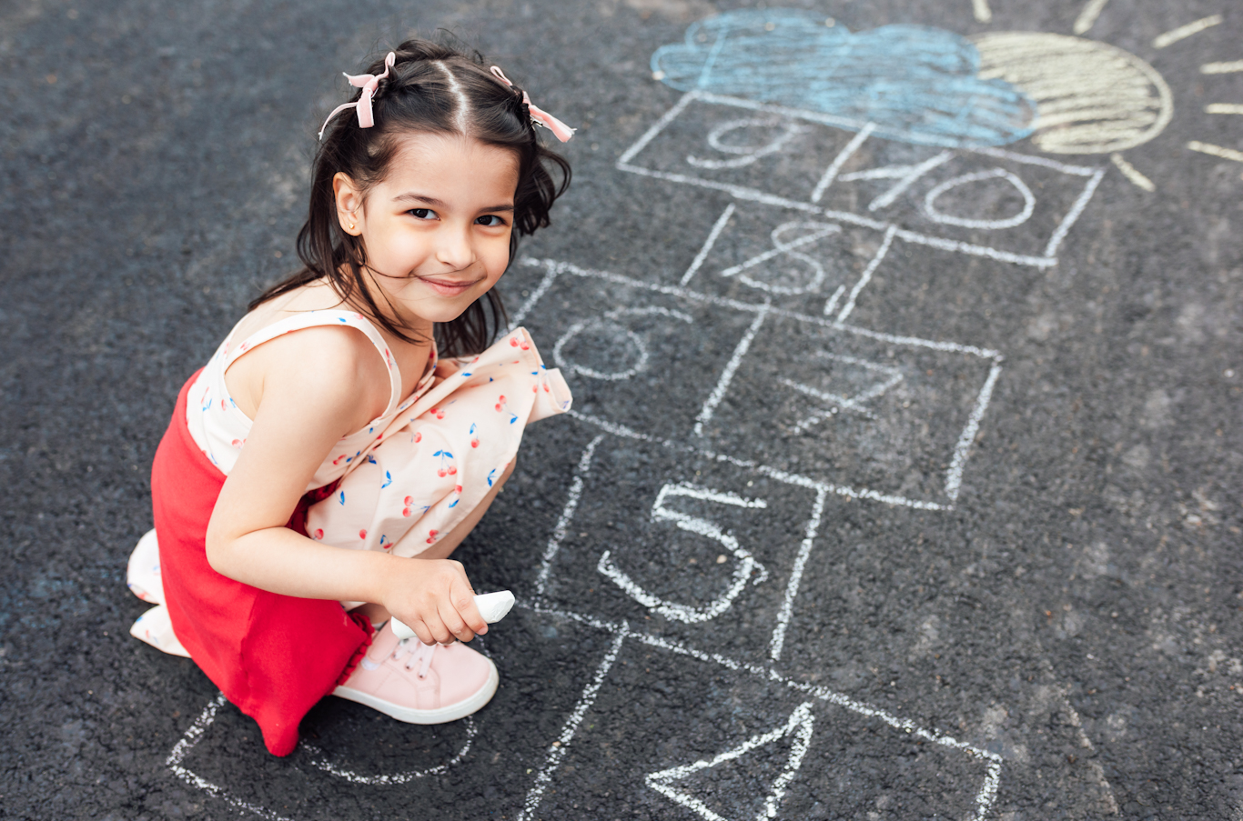 Girl squatting in front of a hopscotch grid drawn on the pavement. The girl is holding a piece of chalk and smiling