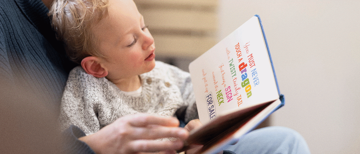 Boy looking at book whilst sitting in his parents lap. The parent is holding the book reading it to the young boy.