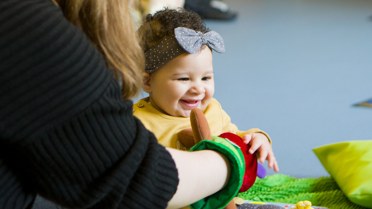 A child giggling with a lady sitting in front of the child with a hand puppet on.