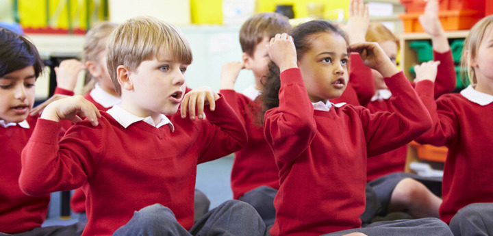Children in classroom wearing school uniforms. The children are sitting cross-legged and singing songs with actions, touching their shoulders