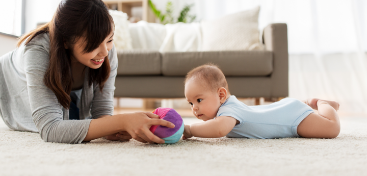 Mother and baby on floor with soft fabric ball. Baby is laying on their stomach on a rug, in a living room.