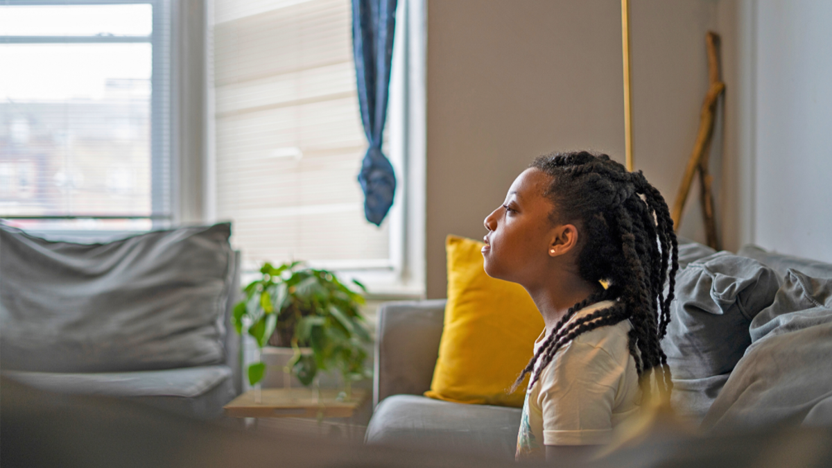Child sat on grey sofa in their living room looking off camera (left) with their child slightly raised.