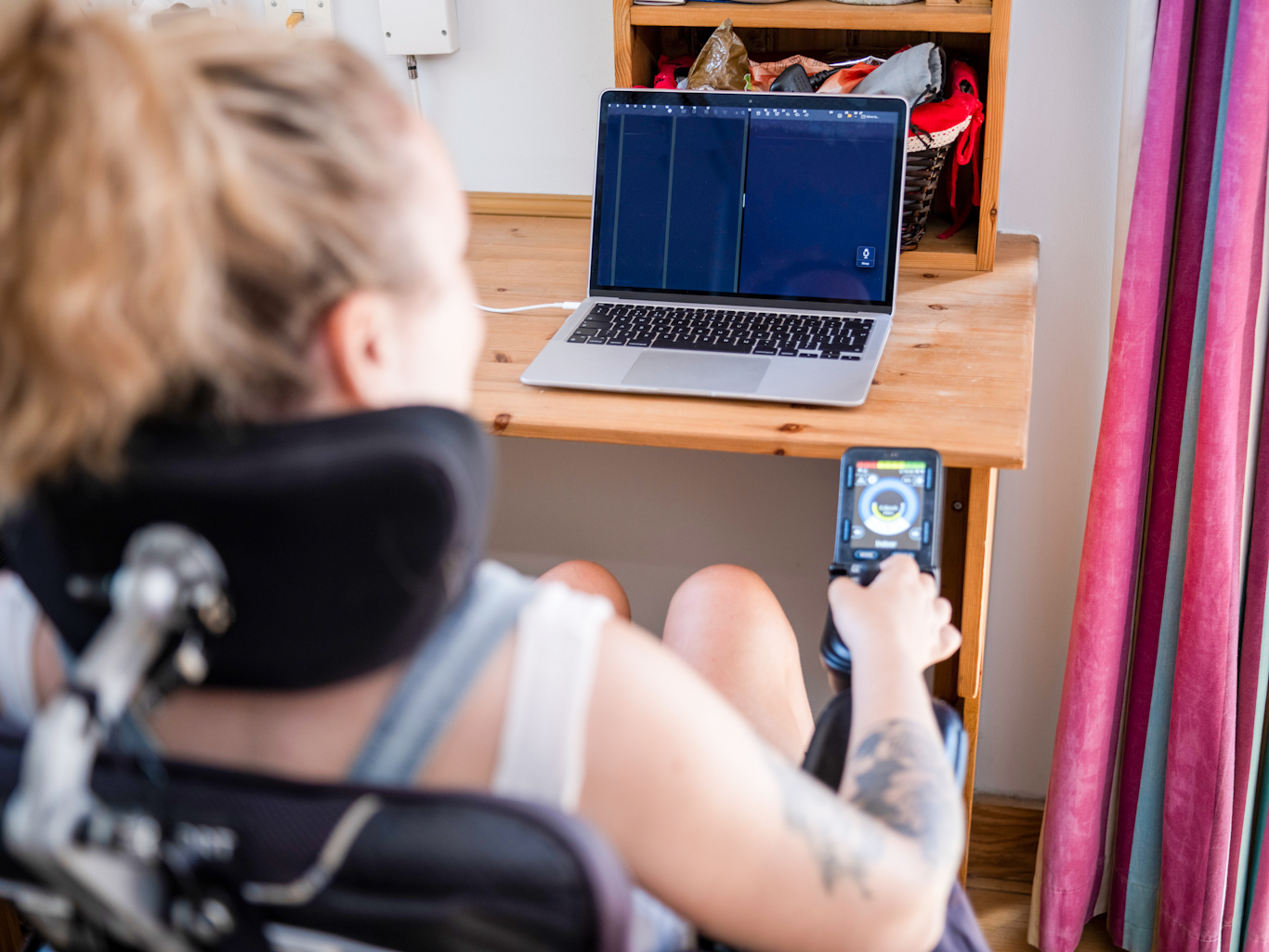Adult woman in wheelchair sitting at desk with her laptop. The focus is on the laptop on the desk and the woman is out of focus in the foreground.